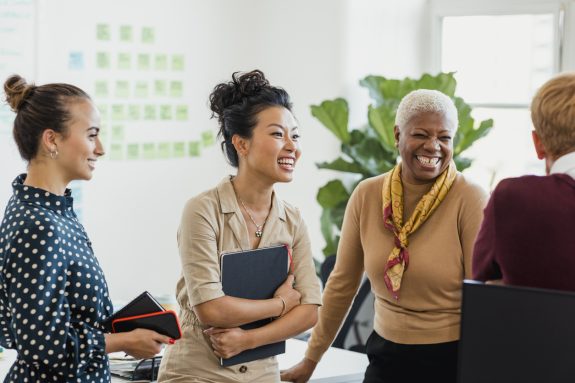 Colleagues standing in a small group discussing something while laughing. Two of the women are holding notebooks.