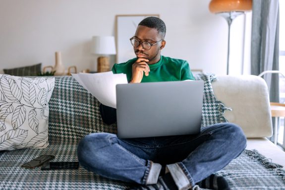 Young casually clothed concentrated man going over paperwork while working from living room, he is sitting on the sofa with laptop in his lap