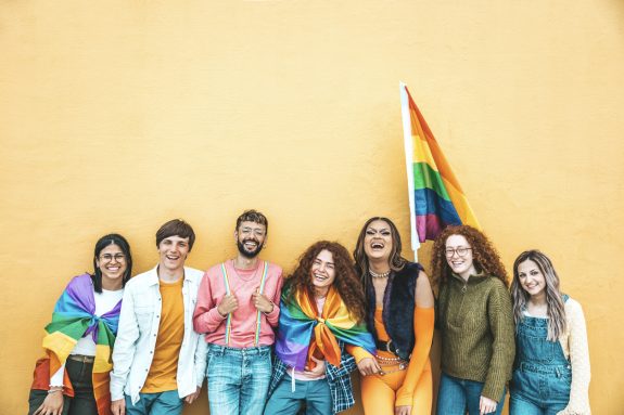 Diverse group of young people celebrating gay pride festival day - Lgbt community concept with guys and girls hugging together outdoors - Multiracial cheerful friends standing on a yellow background